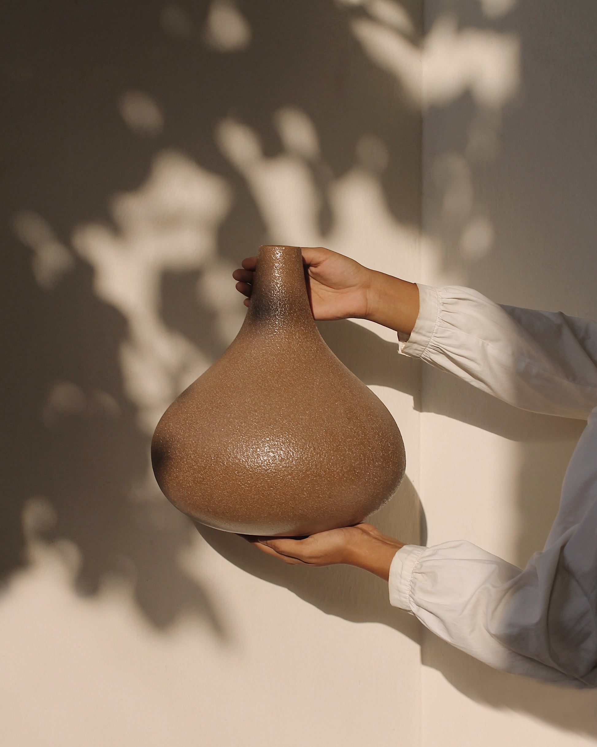 The arms of a woman wearing a white blouse are shown holding a garlic-shaped vase with terracotta and fine black speckles finish sits against a white background with the shadows of a tree branch