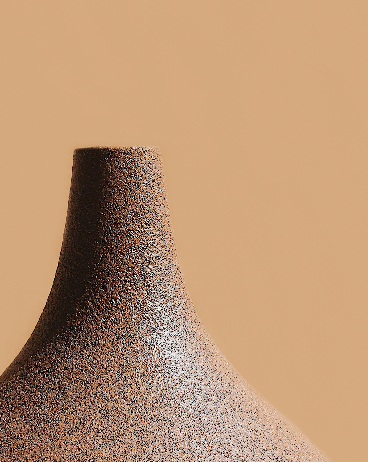 A close-up view of the top of a garlic-shaped vase with a terracotta and fine black speckles finish sitting against a beige background