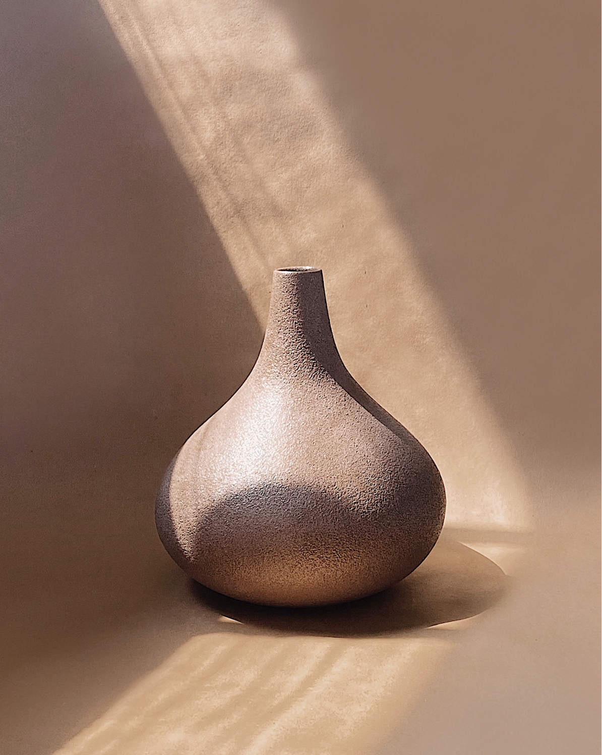 A garlic-shaped vase with a terracotta and fine black speckles finish sits against a beige background with sun rays shining in behind it