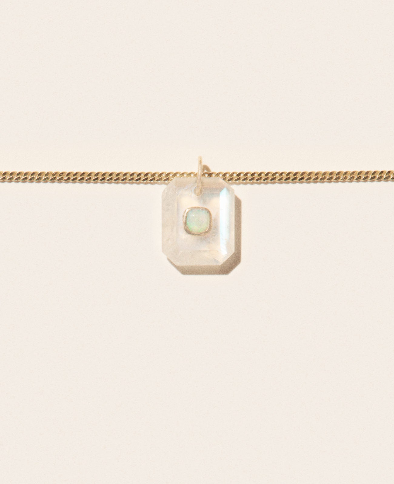 Close up of a segment of a gold chain necklace with a milky moonstone pendant featuring an opal in the center set in yellow gold and sitting on an off-white background