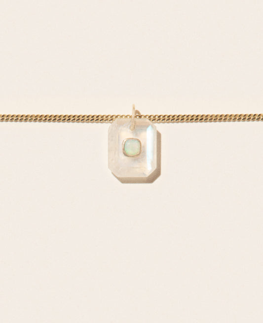 Close up of a segment of a gold chain necklace with a milky moonstone pendant featuring an opal in the center set in yellow gold and sitting on an off-white background