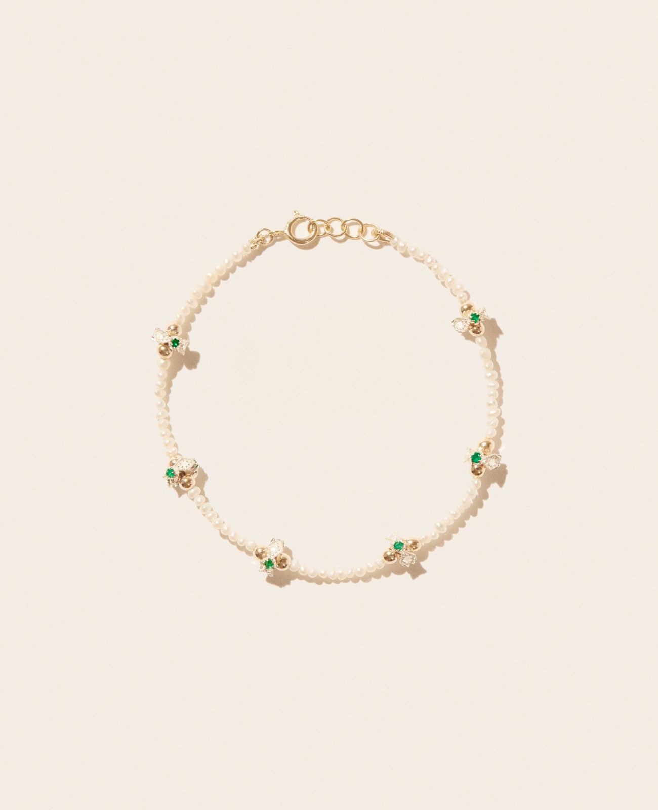 A stringed freshwater pearl bracelet with six clusters of gold beads, diamonds, and emeralds spaced evenly throughout and a gold clasp shown laid flat on an off-white background