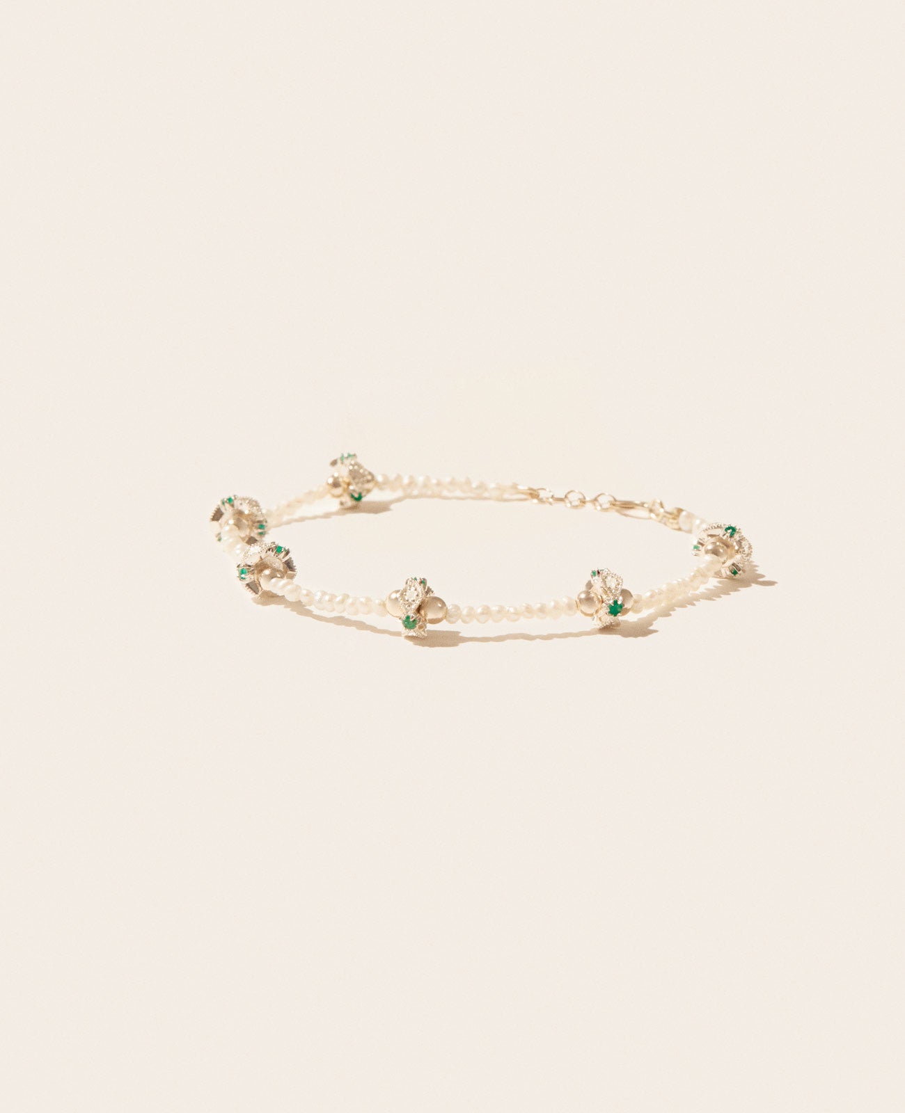 A side view of a stringed freshwater pearl bracelet with six clusters of gold beads, diamonds, and emeralds spaced evenly throughout and a gold clasp shown on an off-white background