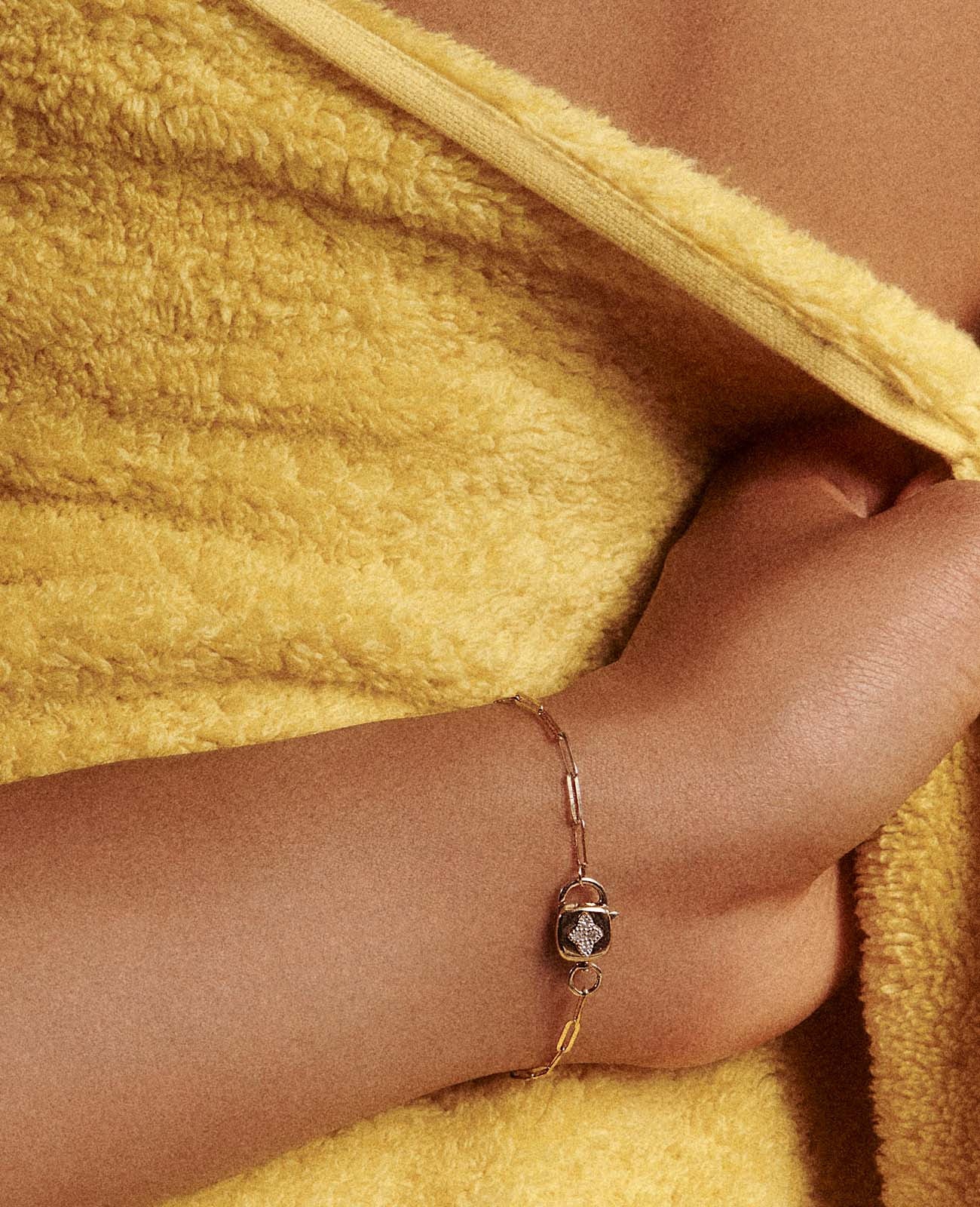 The wrist of a woman in a fuzzy yellow top   is shown with a gold chainlink-style bracelet and a lock-shaped clasp with a diamond four-point star inlay