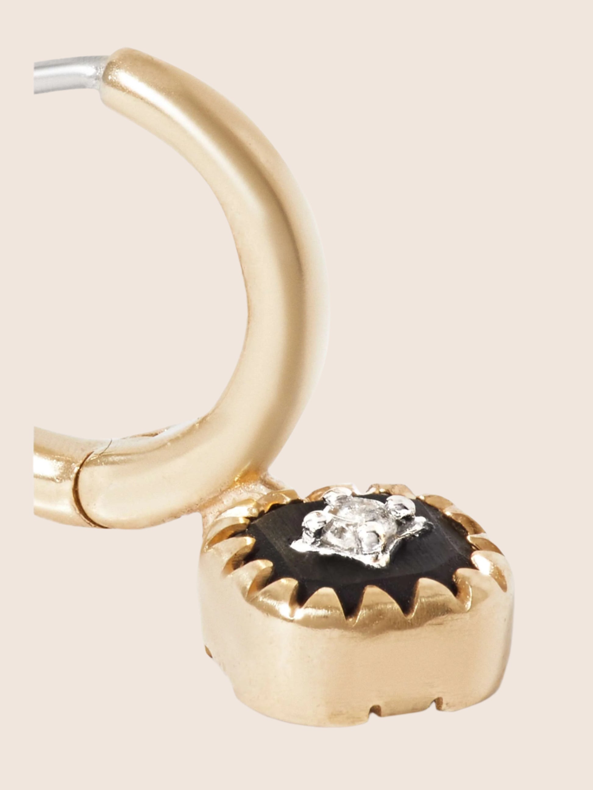 Gold hoop earring with hanging pendant that features a black resin medallion with a diamond placed in the center and set in gold with a sun ray design encircling the front design resting on its back on an off-white surface