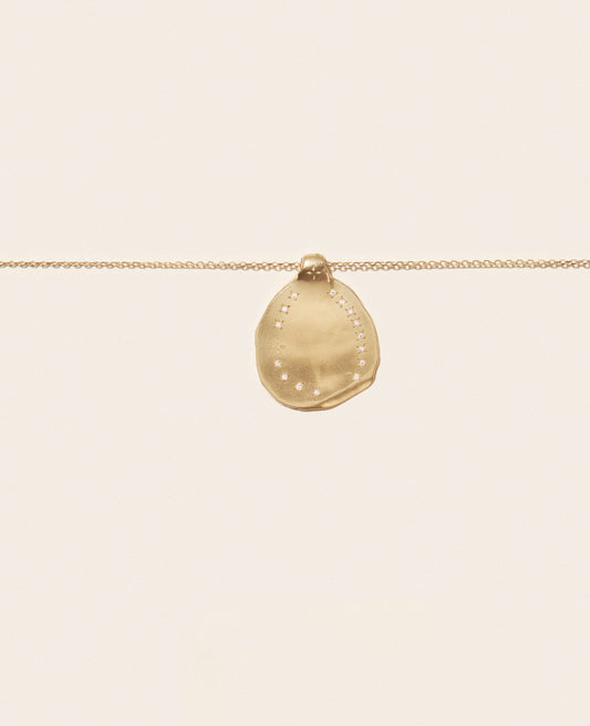 A close up view of a yellow gold irregular shaped round pendant with ring of inlaid diamonds set on a gold chain necklace with an off-white background