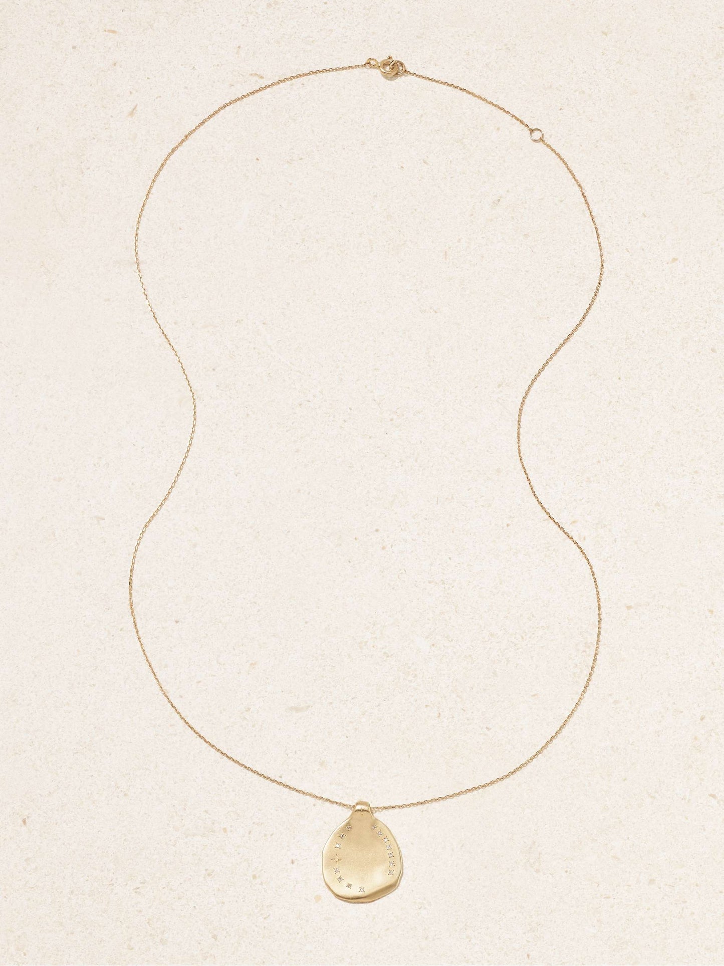 A yellow gold irregular shaped round pendant with ring of inlaid diamonds set on a gold chain necklace with an off-white background
