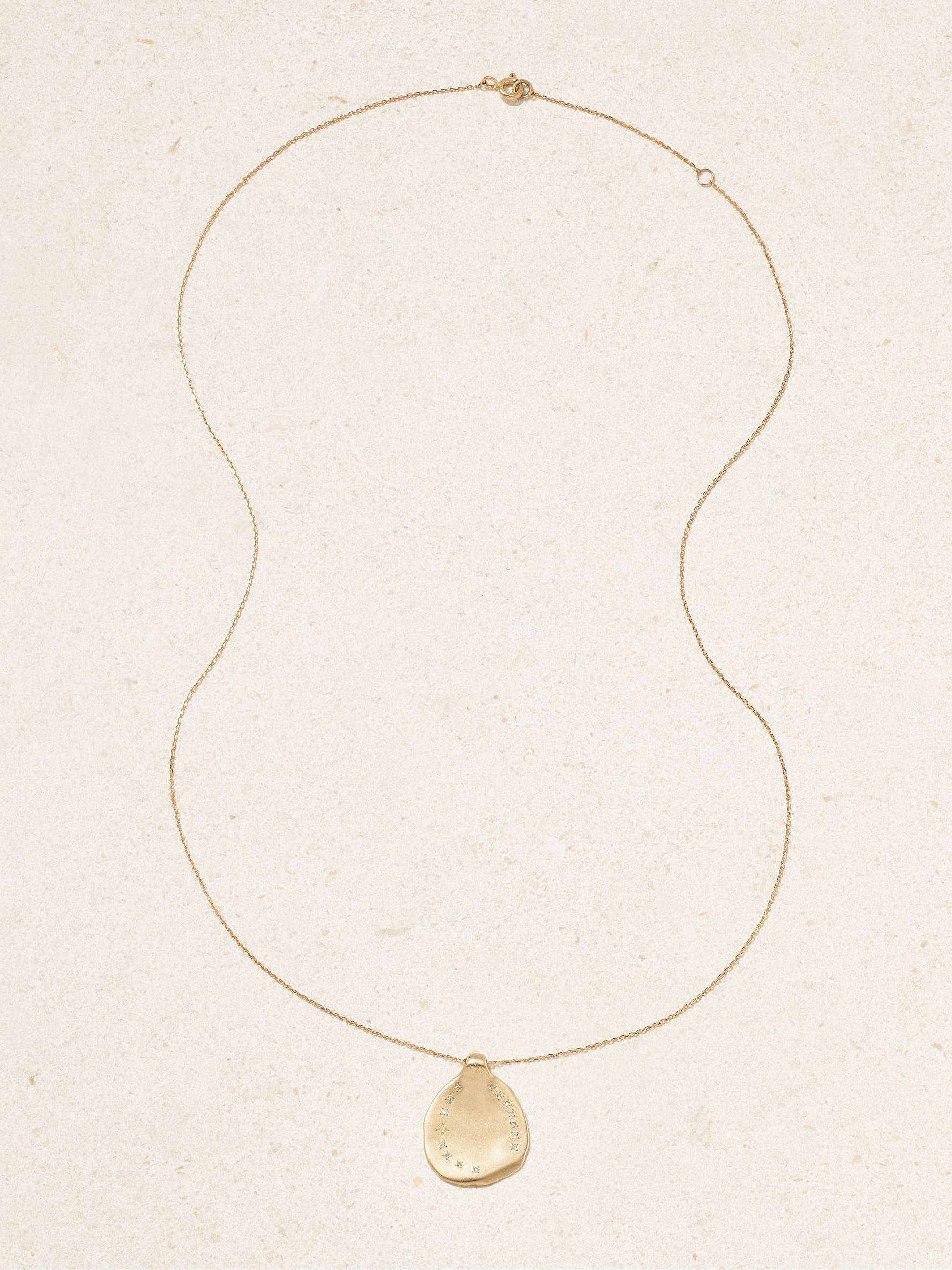 A yellow gold irregular shaped round pendant with ring of inlaid diamonds set on a gold chain necklace with an off-white background