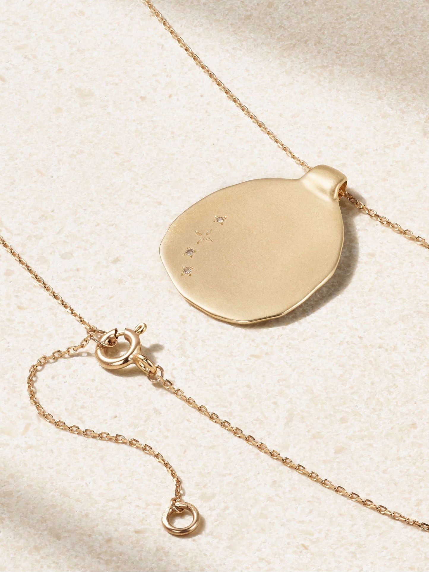 A closeup of the back side of a yellow gold irregular shaped round pendant with string of inlaid diamonds set on a yellow gold chain necklace with an off-white background