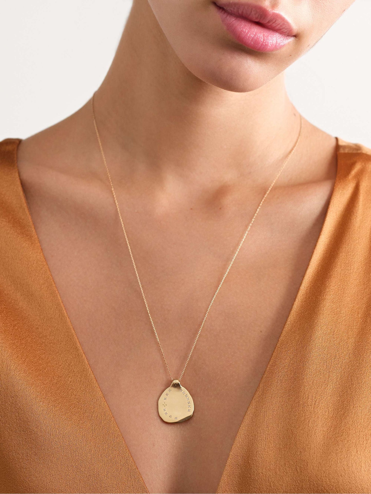 A view of the neck and chest of a woman in a bronze-colored v-neck blouse wearing a yellow gold necklace with an irregular shaped gold round pendant with ring of inlaid diamonds 