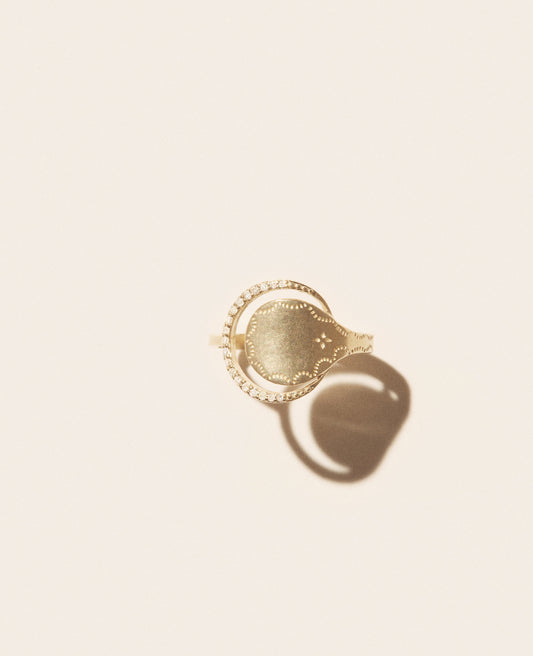 A gold ring with a round shape tucked into a half-ring of diamonds set against an off-white background shown from the top view