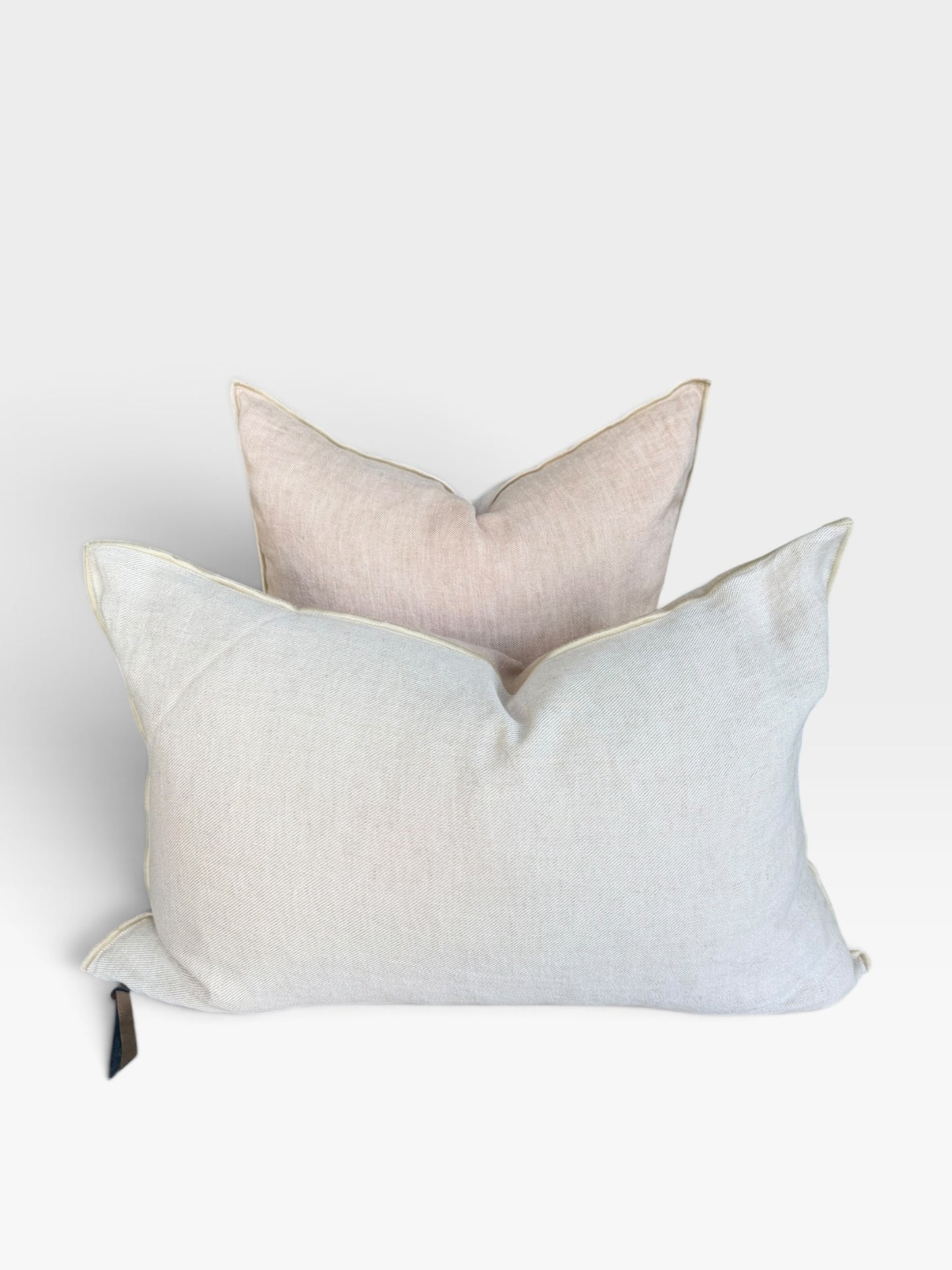 Two square decorative throw pillows stacked together on a light gray background. The front pillow is a light beige with a small black leather tie at the lower left, and the pillow behind it is a deeper sandy beige.