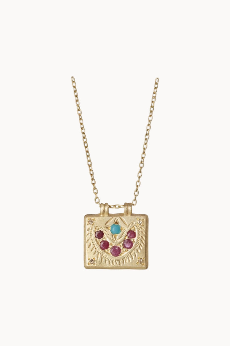 A gold necklace with a square gold charm adorned with rubies, turquoise and diamonds hangs against a soft white background