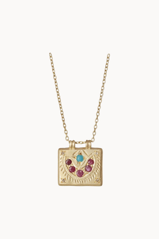A gold necklace with a square gold charm adorned with rubies, turquoise and diamonds hangs against a soft white background