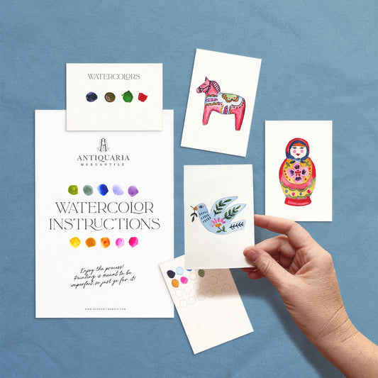 A hand holds a printed watercolor instruction card from Antiquaria, surrounded by other printed cards and swatches on a blue textured background. The cards feature Scandinavian folk art images, including a pink horse, a colorful matryoshka doll, and a blue bird with green leaves.