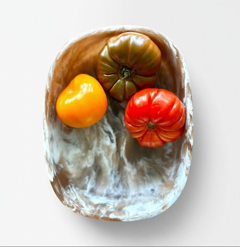 A top-down view of a rustic, oval-shaped ceramic resin bowl with a marbled brown, white, and gray coloration containing three heirloom tomatoes (one green, one red, one orange-yellow) on a light gray background.