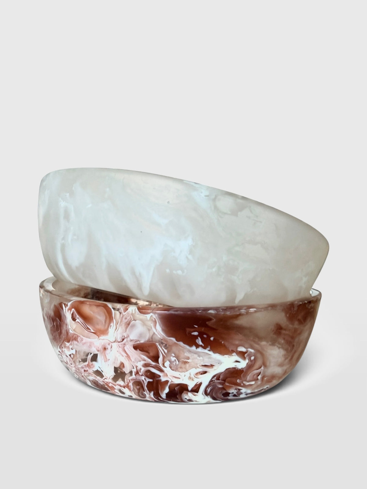 Two stacked round resin bowls on a light gray background. The top bowl is milky translucent white, and the bottom bowl has a marbled swirl pattern of brown, red, pink, and white.