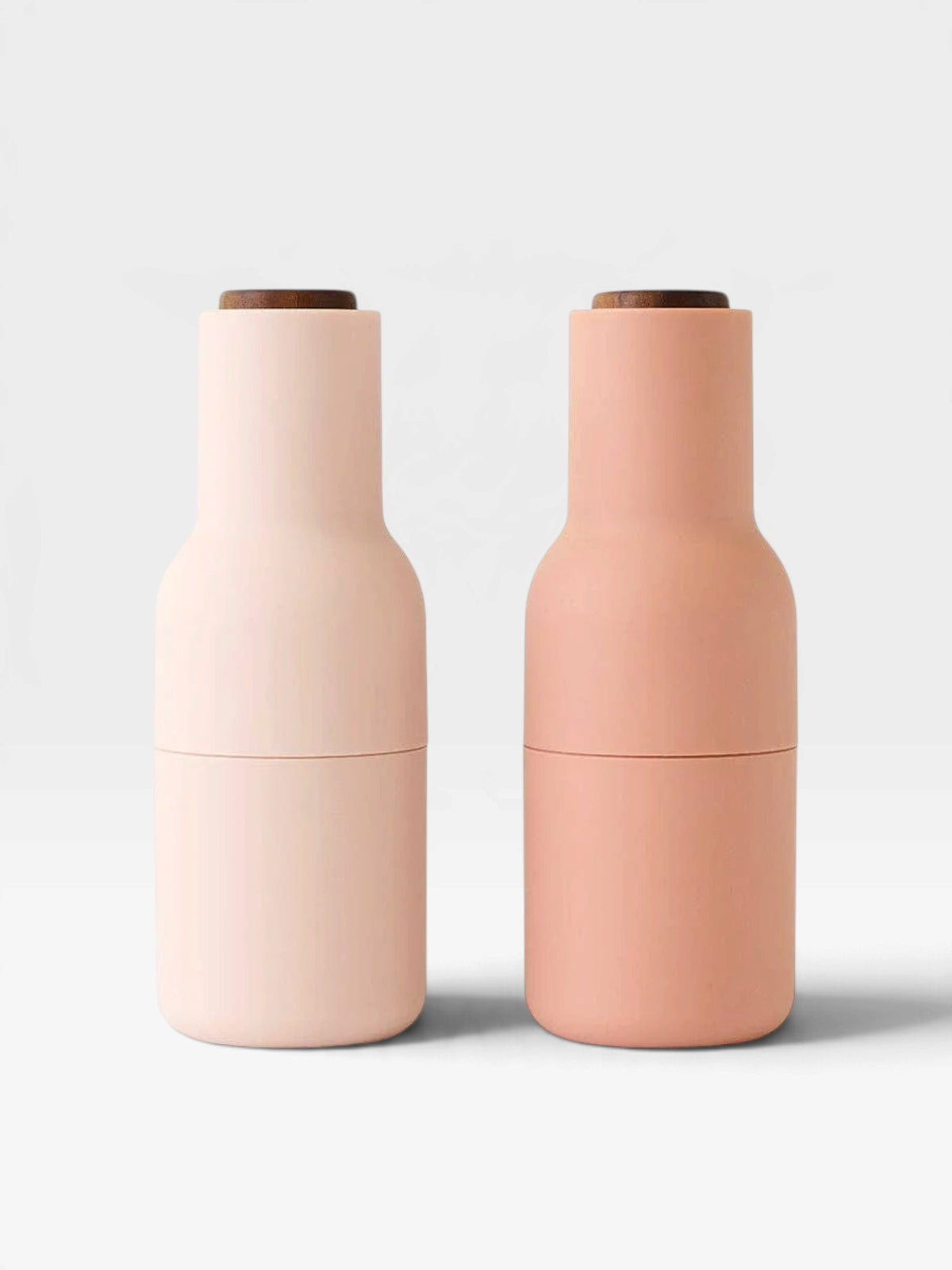Two matte bottle-shaped containers standing side by side on a light gray background. The left container is a very pale blush pink, and the right container is a soft peachy pink.