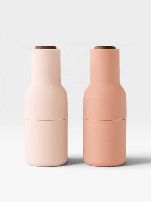 Two matte bottle-shaped containers standing side by side on a light gray background. The left container is a very pale blush pink, and the right container is a soft peachy pink.