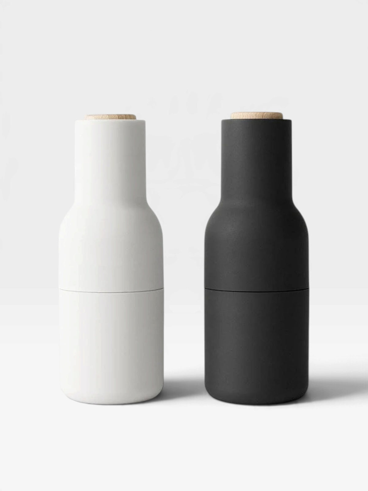 Two spice bottle grinders, one in white (Ash) and one in black (Carbon), with beech wood lids on a white background