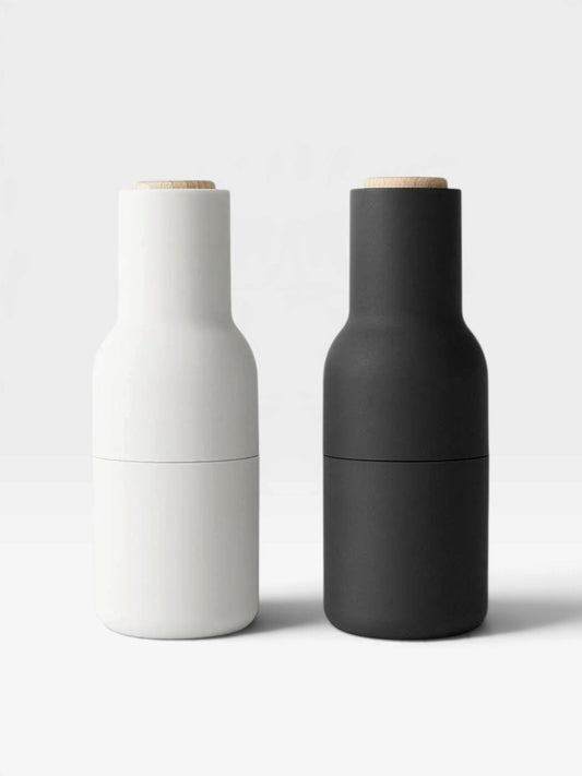 Two spice bottle grinders, one in white (Ash) and one in black (Carbon), with beech wood lids on a white background