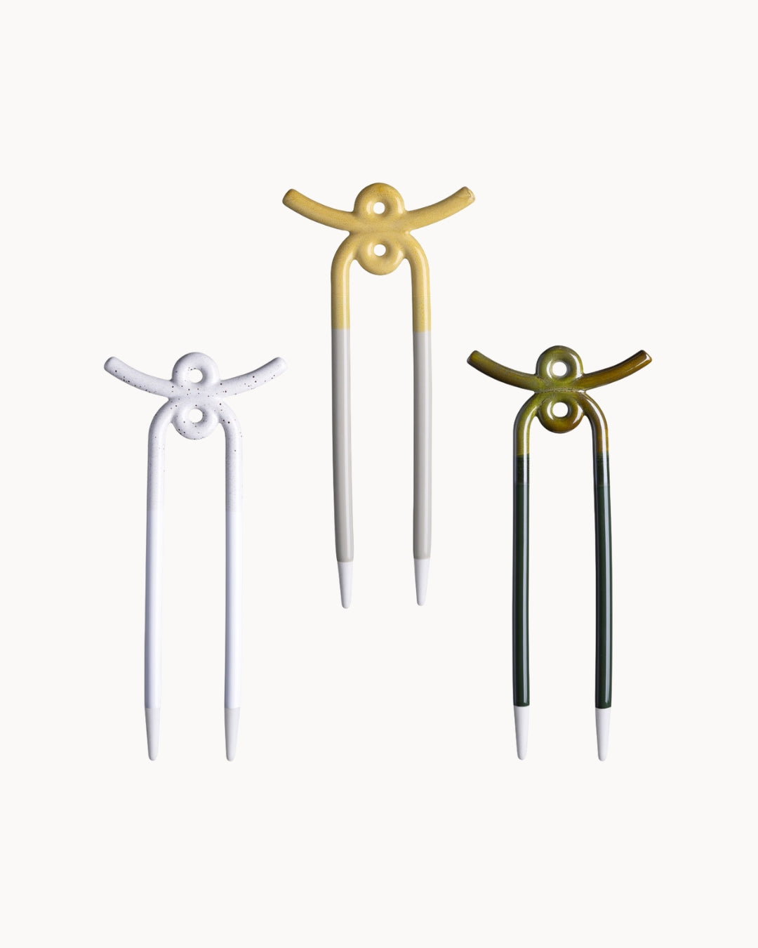 Three ceramic plant stakes shaped like stylized figures, shown on a plain light background. The left pin is white, the center pin is white with a gold top, and the right pin is dark green with a gold top.