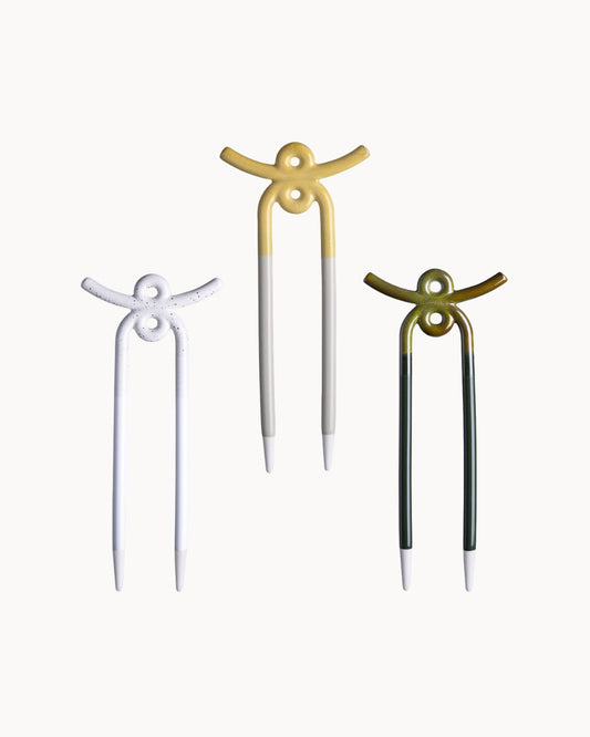 Three ceramic plant stakes shaped like stylized figures, shown on a plain light background. The left pin is white, the center pin is white with a gold top, and the right pin is dark green with a gold top.