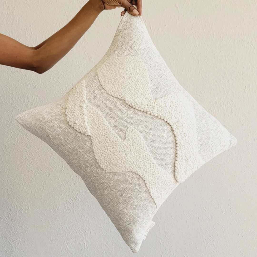 Textured ivory pillow with knitted wave design held by a hand against a neutral background