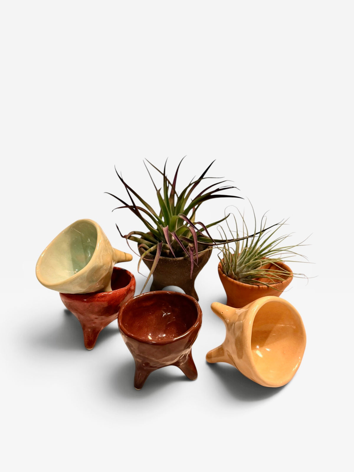 A set of six handcrafted ceramic mini planters in various earthy colors, including terracotta, brown, orange, and pale yellow, some of which are stacked and others holding small potted air plants, photographed on a clean white background.
