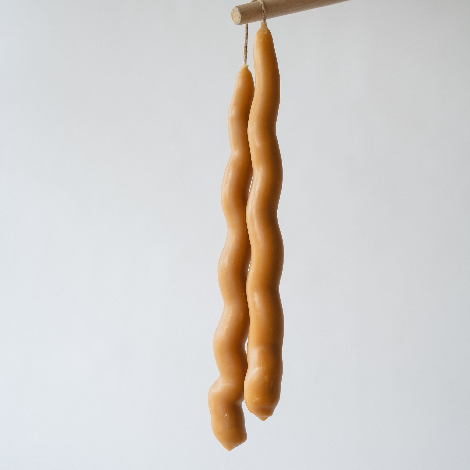 A pair of wavy taper candles made of beeswax, hanging against a light background.