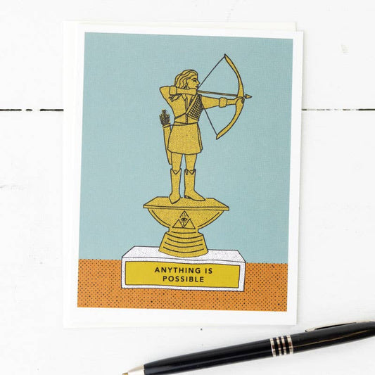 A greeting card with a illustration of a figure with a bow and arrow on a pedestal with the text 'Anything Is Possible' on the base, placed on a white surface with a pen beside it.