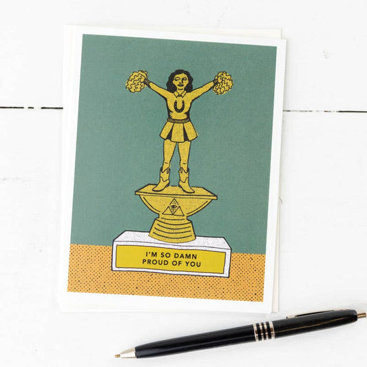 A greeting card with a cartoon character holding pom poms and wearing a crown, with the text 'I'm So Damn Proud of You' on it. The background is green and there is a pen and a stamp beside the card.