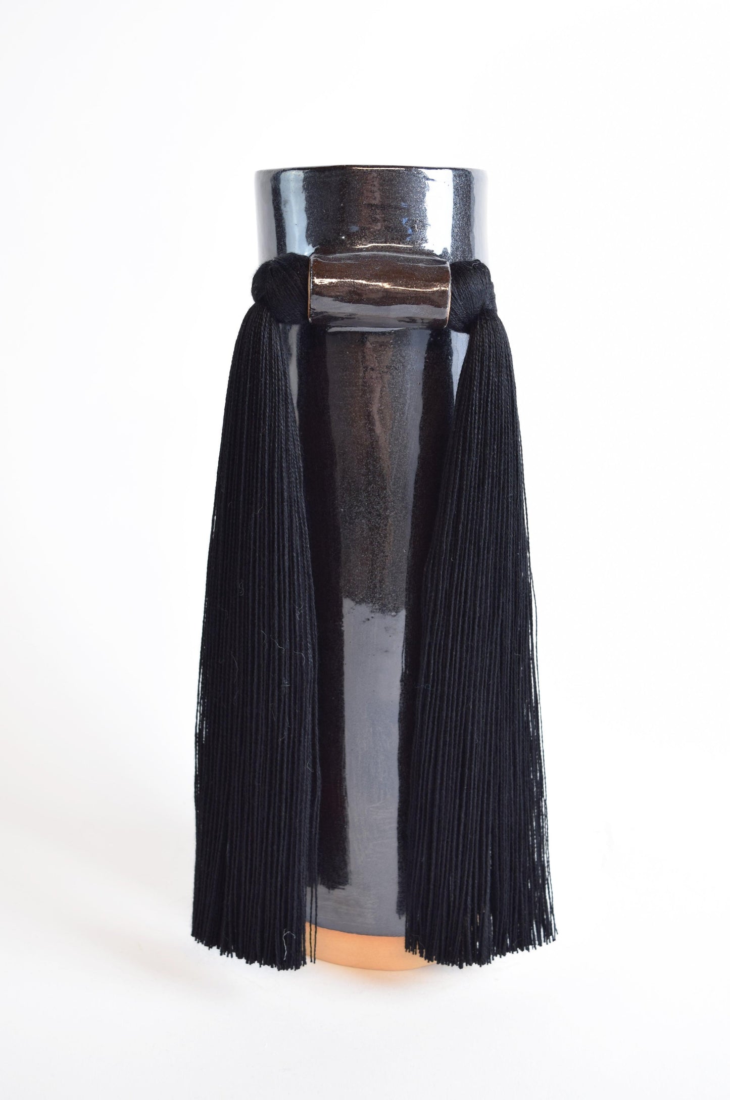 A black stoneware vase with smooth cotton fringe hanging from the rim, displayed against a white background.