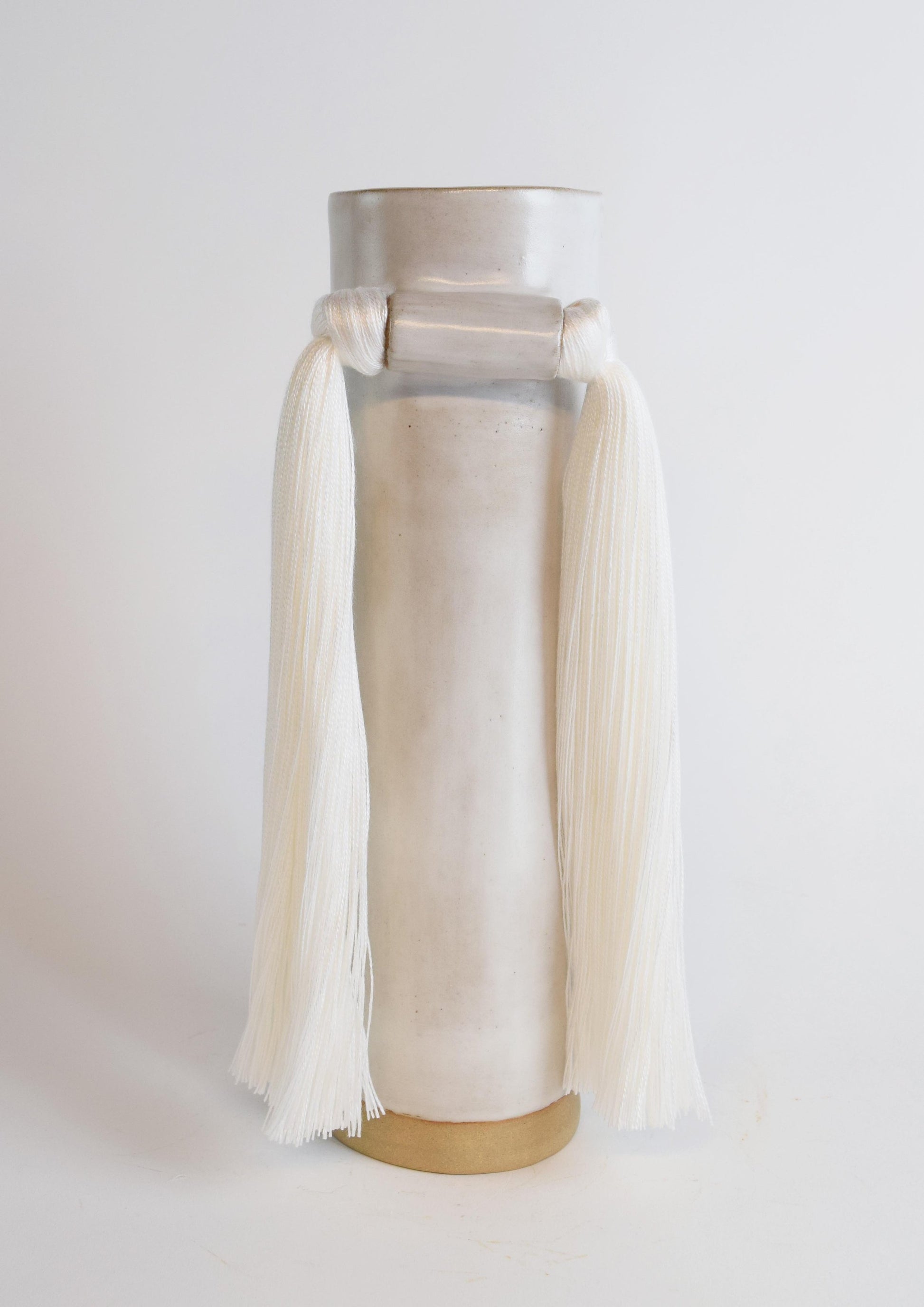 A white stoneware vase with ivory cotton fringe hanging from the neck, displayed against a neutral background.