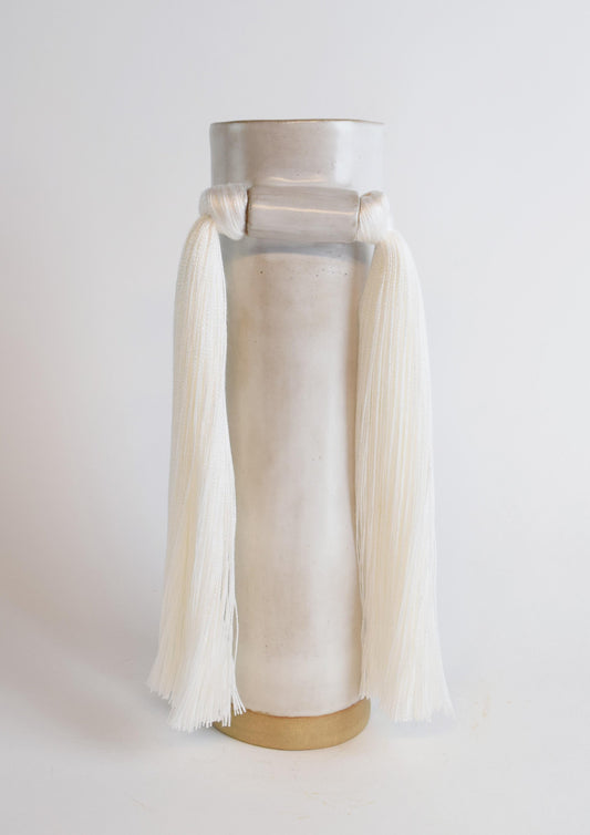 A white stoneware vase with ivory cotton fringe hanging from the neck, displayed against a neutral background.