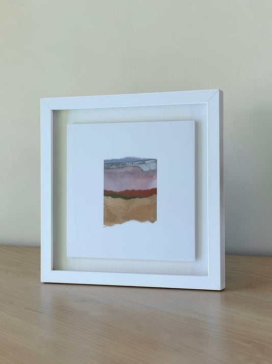 A white square shadow box frame displaying a small abstract landscape art print with layered pink, red, green, and tan paper textures, set on a light wood surface against a neutral beige wall.