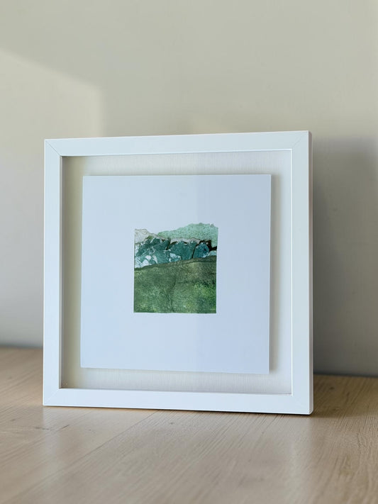 A square white-framed art print leaning against a beige wall on a light wood surface. The print features a small textured green and blue landscape artwork centered on a white mat.