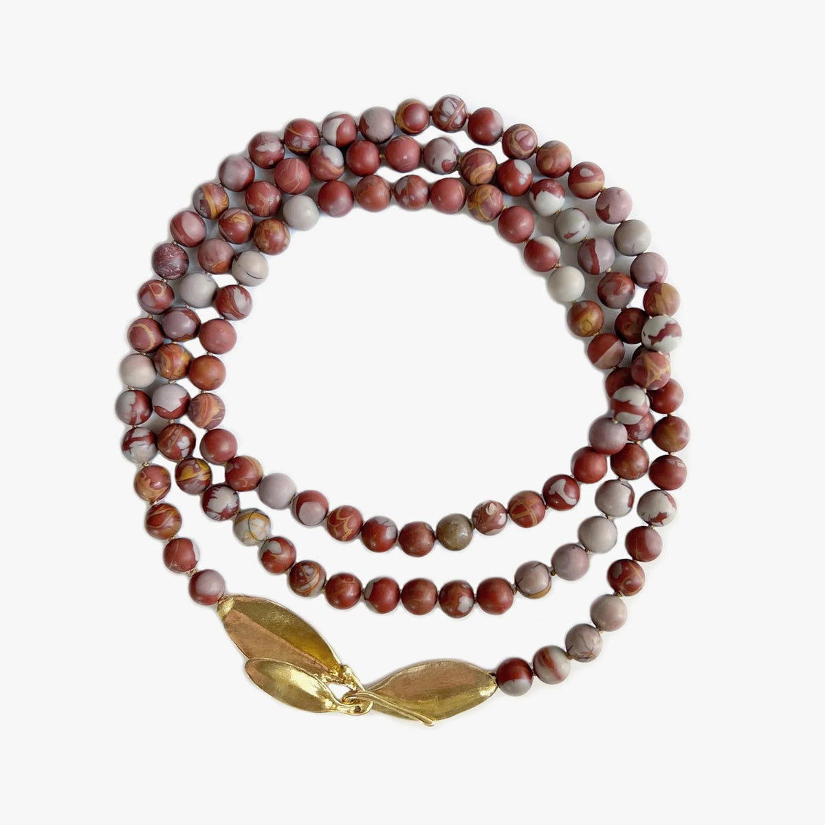 A necklace made of jasper beads in deep red and soft grays and a leaf-shaped clasp sits coiled in three rings on a light gray background.