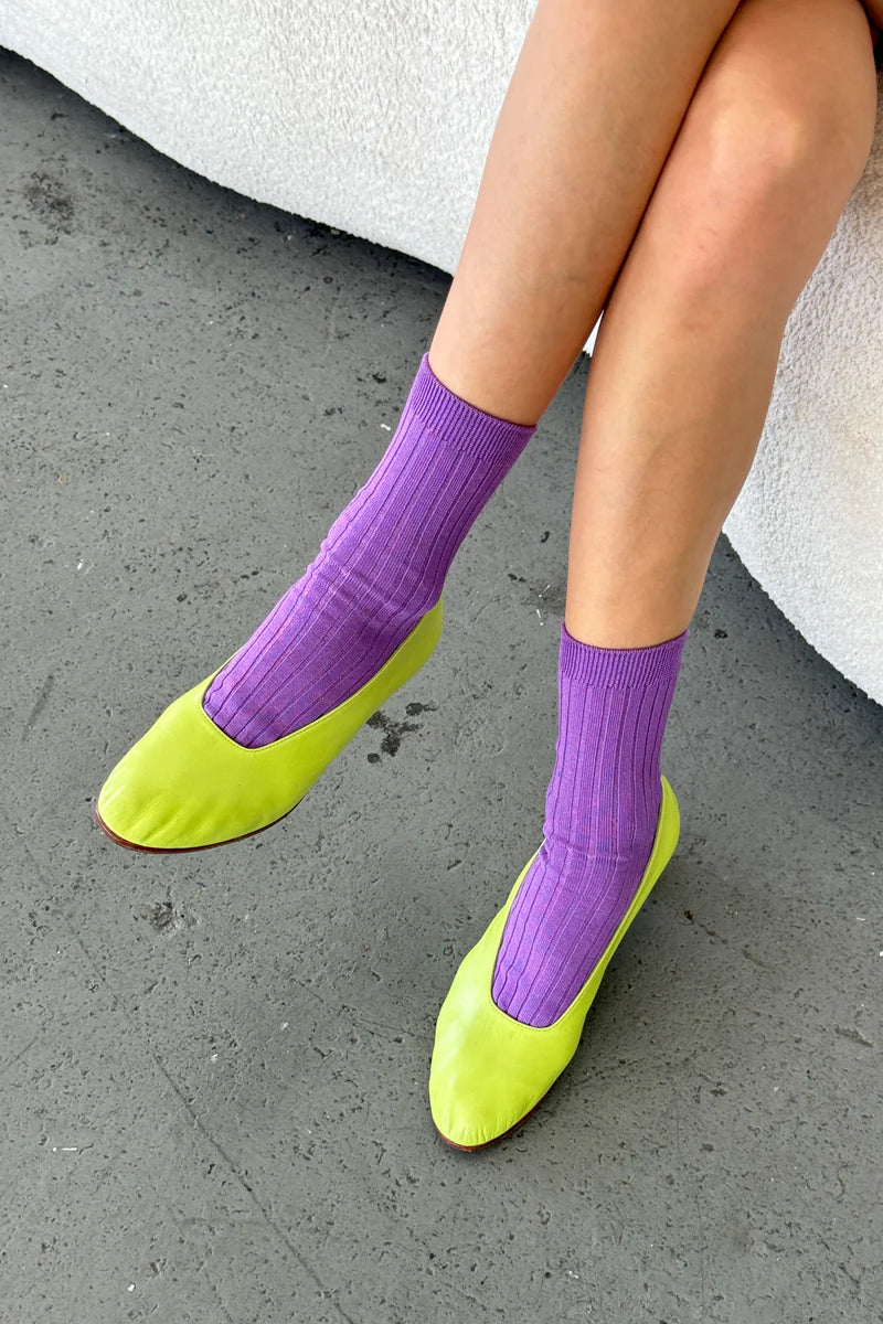 Person wearing purple le bon shoppe socks and lime green shoes on a concrete surface.