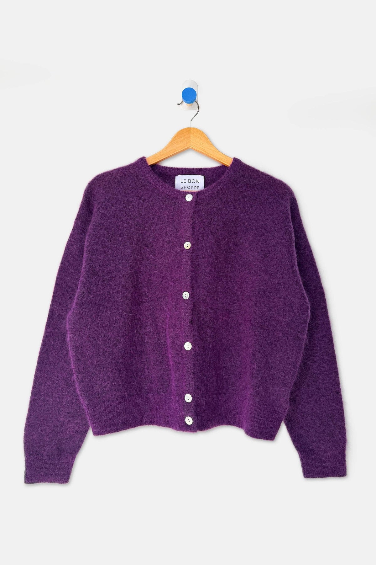 A purple knit cardigan sweater with white buttons, hanging on a wooden hanger against a light gray background.