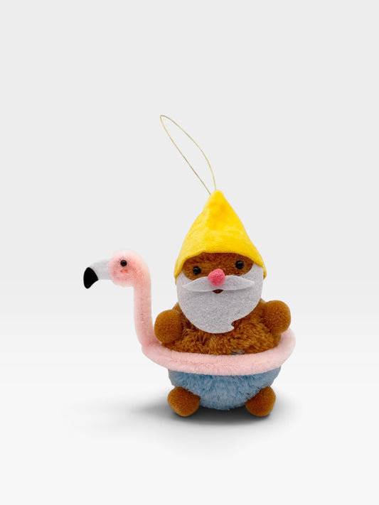 A plush gnome ornament sitting in a pink flamingo-shaped bathtub, wearing a yellow hat and blue pants, photographed on a clean white background.