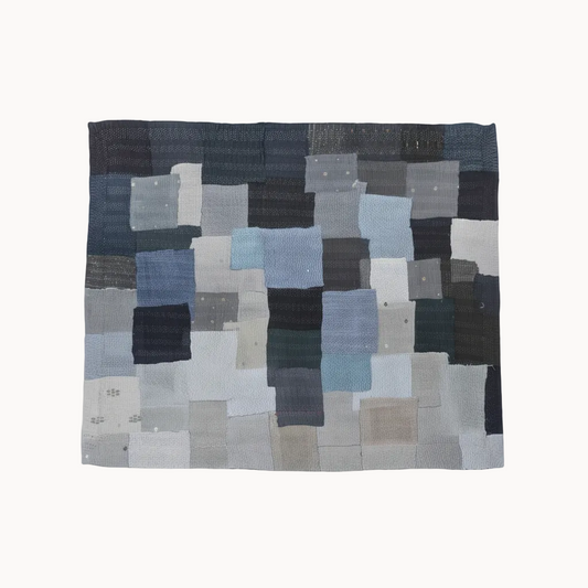 A square textile wall hanging or quilt made from patchwork fabric squares in muted blue, gray, charcoal, beige, and off-white tones, photographed against a plain white background.