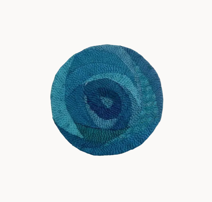 A round woven rug with a textured spiral pattern in various shades of blue and teal, shown against a plain white background.