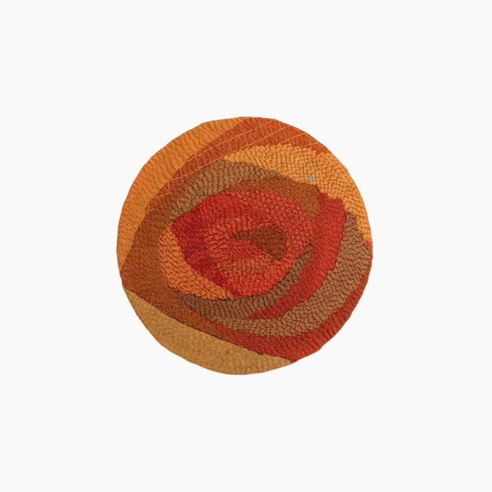 A round woven rug with a textured, layered abstract pattern in shades of orange, red, and brown, centered on a light gray background.