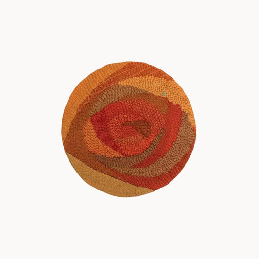 A round woven rug with a textured, layered abstract pattern in shades of orange, red, and brown, centered on a light gray background.