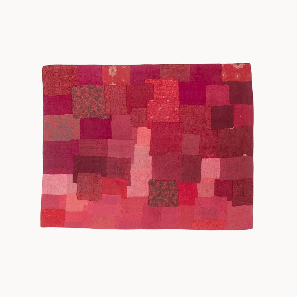 A square patchwork textile artwork made of many red, pink, and burgundy fabric squares, shown flat against a plain white background.