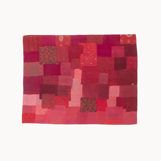 A square patchwork textile artwork made of many red, pink, and burgundy fabric squares, shown flat against a plain white background.