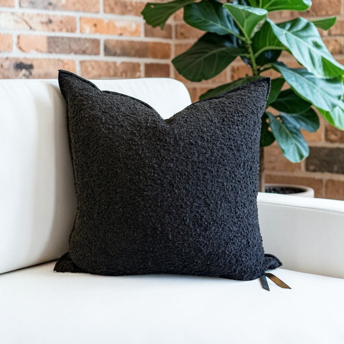 A square black textured throw pillow with a bouclé-like fabric, resting on a white sofa in front of a brick wall and a green potted plant.