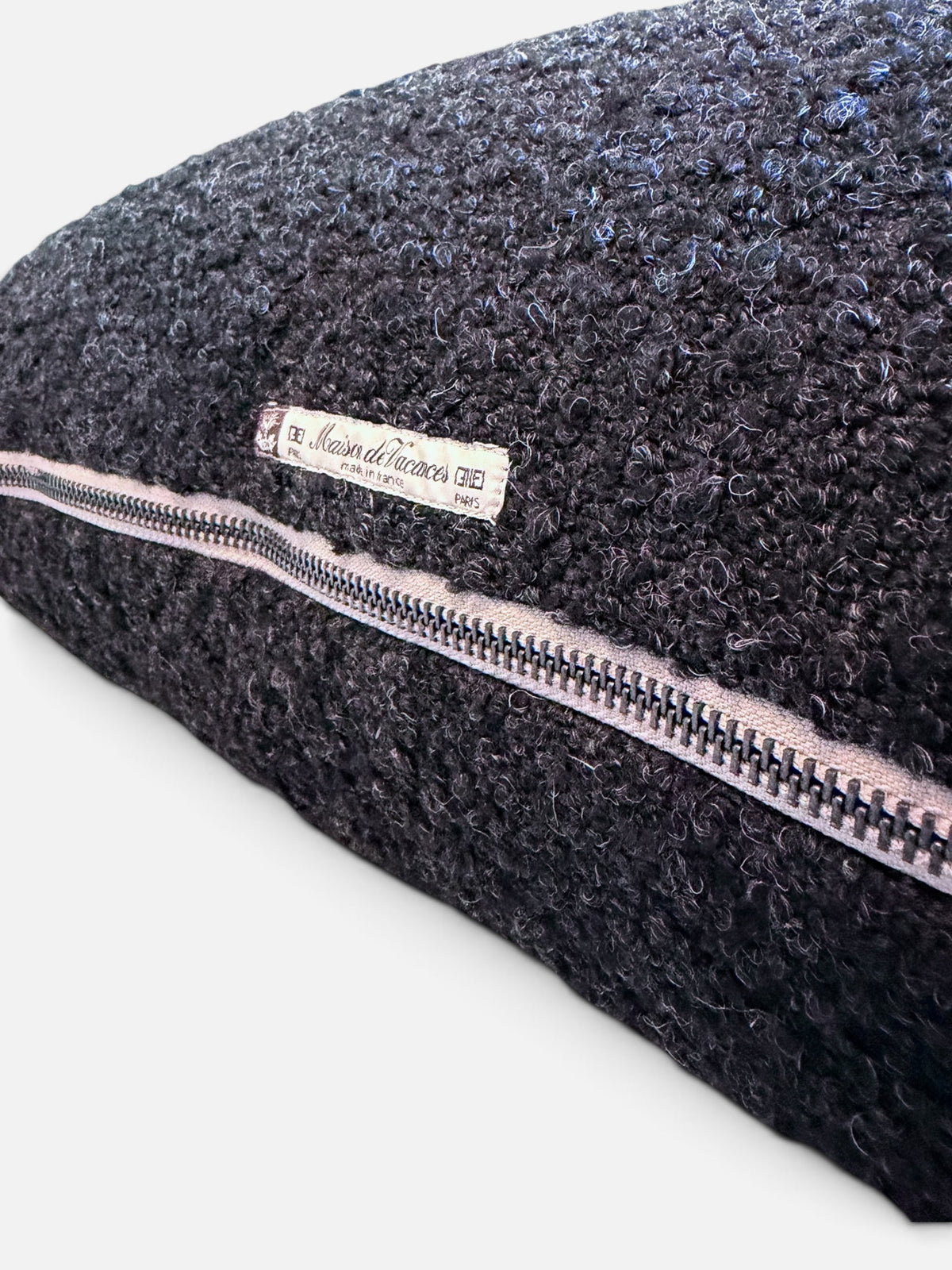 Close-up of a dark charcoal gray bouclé pillow with a light pink zipper trim and a white brand label reading 'Maison de Vacances'.