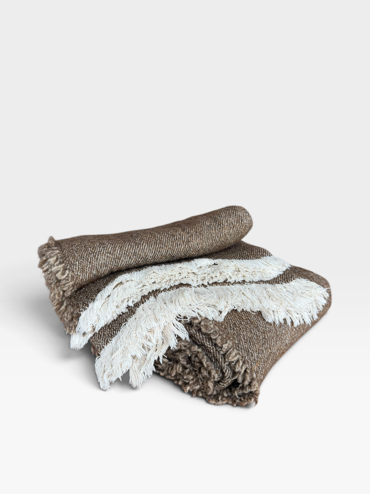 A folded woven throw blanket with a brown and white striped pattern and fringed edges, photographed on a clean light gray studio background.